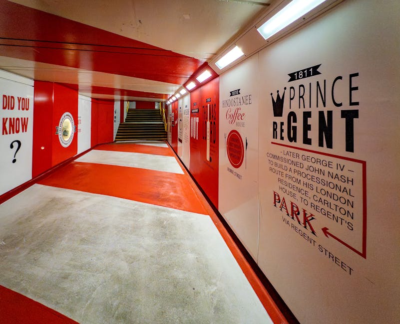 A brightly lit underground corridor with polished concrete flooring featuring a red-painted central strip and light grey sides, leading to a staircase with dark, non-slip treads. The walls are decorated with large, vintage-style advertisements and informational posters in black, red, and white, including a prominent sign reading 'Prince Regent' with historical details. The ceiling is fitted with white lighting fixtures providing even illumination, and the overall scene appears clean and well-maintained, exemplifying surface cleaning and sanitisation in a commercial or public space. The setting reflects a focus on hygiene and upkeep aligned with the services offered by Marylebone Carpet Cleaners.