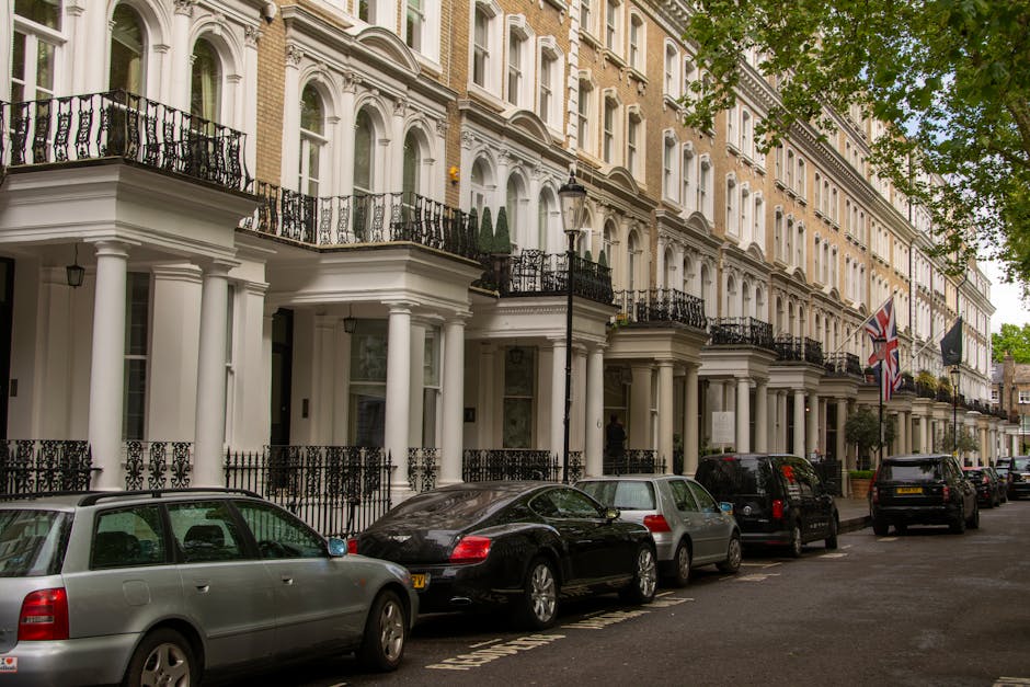 A row of elegant, white Victorian-style residential buildings with decorative columns and intricate black wrought-iron balconies lining a tree-lined street in Marylebone, London. Several parked cars, including a silver station wagon, a black sedan, and a grey hatchback, are positioned along the curb. The buildings are adorned with Union Jack flags and street lamps, and the scene is illuminated by natural daylight with lush green foliage from trees overhead, creating a tidy and well-maintained urban setting. This image showcases the characteristic architecture of Marylebone, emphasizing cleanliness and preservation typical of high-end residential areas, relevant to deep cleaning and surface maintenance in the local context.