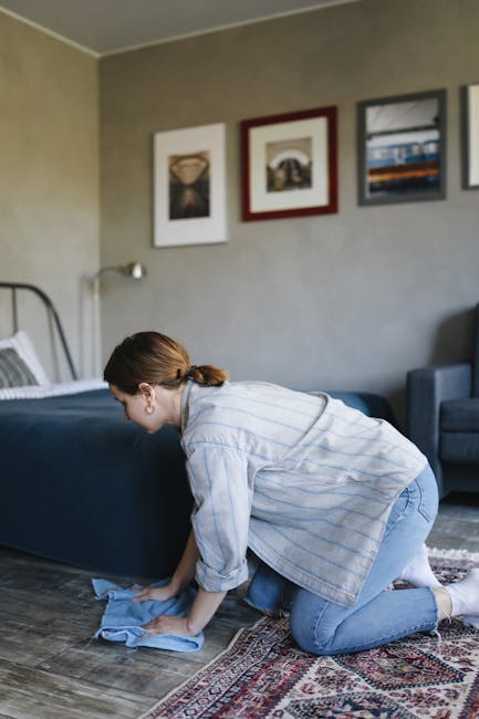 A professional cleaning technician dressed in a full white protective suit, gloves, and a face mask is performing deep carpet cleaning in a bright, minimalist living room. The technician uses a portab