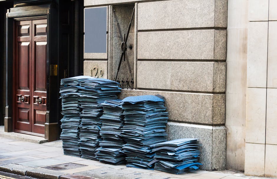 A pile of neatly stacked blue and white printed paper documents situated on the sidewalk outside a building with a stone and brick facade. The documents are organized in several tall stacks beside a b