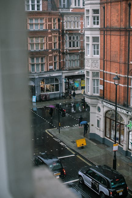A view of a busy street scene captured from a high window, showing wet asphalt pavement with parked cars and pedestrians walking with umbrellas on a rainy day. The street is lined with red-brick and white-painted buildings, including a medical clinic and various shops. The overcast sky reflects on the shiny, damp surfaces, and streetlights are illuminated. The scene depicts urban cleanliness and the need for surface cleaning and maintenance, typical of a central London area such as Marylebone, aligning with the content on Marylebone Carpet Cleaners' website about surface cleaning and hygiene upkeep in residential or commercial settings.